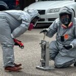 Law enforcement in hazmat suits bent over a many pill press outside on the street.