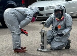 Law enforcement in hazmat suits bent over a many pill press outside on the street.