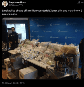 A twitter post showing a police officer standing at the far end of a table with bags of pills and other drug supplies on it. A pill press is visible in the foreground.